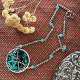 A Sterling silver necklace with natural turquoise blue-green stone inlay and small turquoise beads, arranged on a textured rust-red fabric alongside decorative silver mirror and dried flowers.